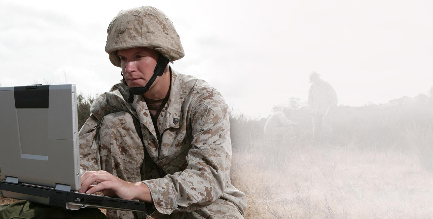 一名身穿战斗装备的男子跪在战场上，在军用笔记本电脑上打字 