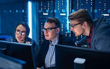 Two men and a woman, all wearing glasses, utilizing Viasat software on computers in a server room