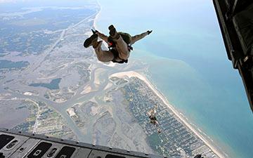 View from an open plane door of two military personnel skydiving towards a coastline