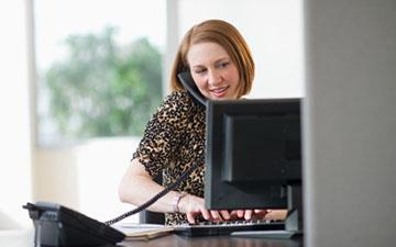 Business woman sitting behind a computer monitor at a desk, typing on a keyboard and talking on the phone